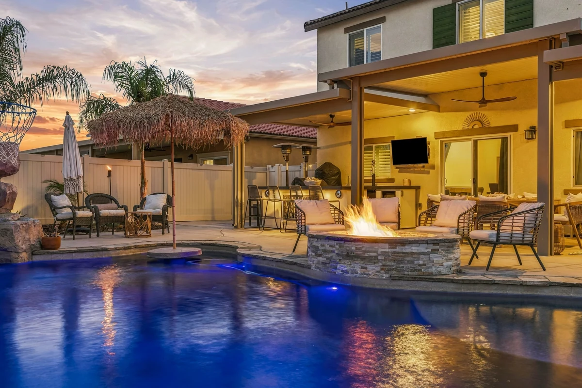 Blue-lit pool beside a covered patio with seating and a fire feature