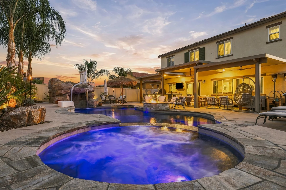 Illuminated spa and curved pool in front of a two-story home at dusk