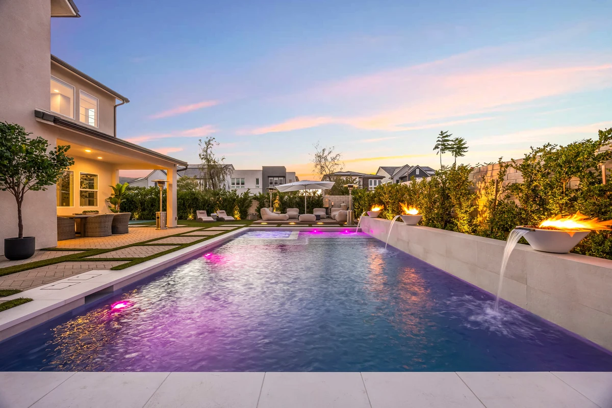 Long backyard pool with colored lighting, fire bowls and an evening sky
