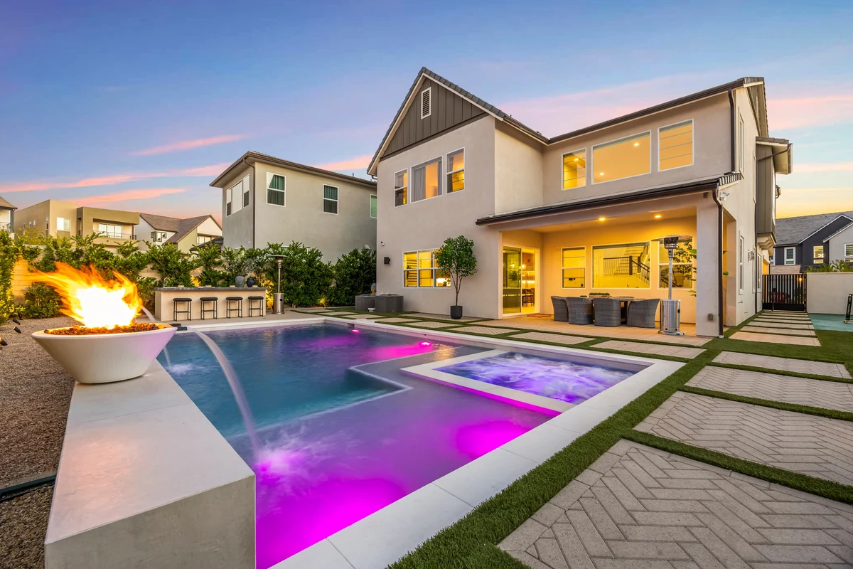Two-story home beside a rectangular pool with colored lighting and a fire bowl at dusk