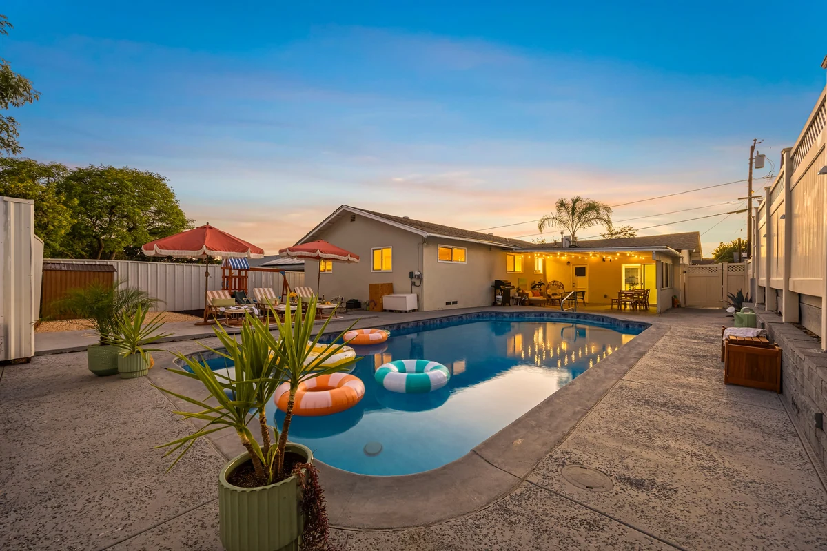 Backyard pool at dusk with an illuminated single-story home and red patio umbrellas