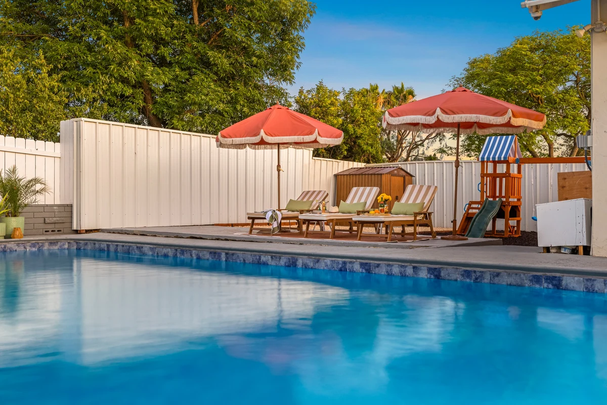 Low view across blue pool water toward loungers and a white fence